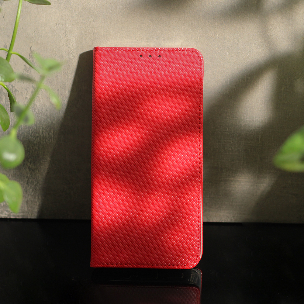 Czerwone etui na telefon z teksturą, stojące na ciemnej powierzchni, w tle widoczne są liście roślin.