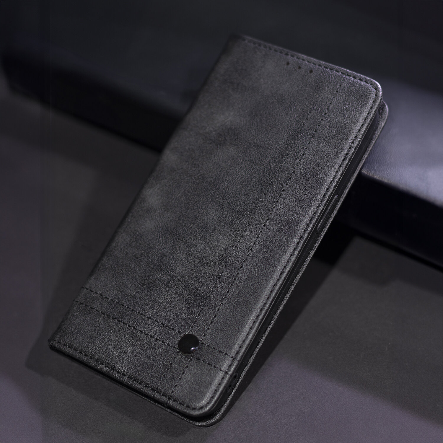 Czarne etui na telefon z widocznymi przeszyciami i małym, okrągłym elementem dekoracyjnym. Etui jest zamknięte i leży na ciemnym tle.