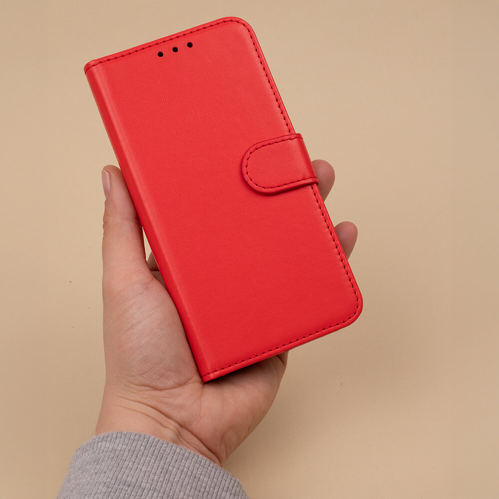 Osoba trzyma czerwone etui na telefon z klapką, wykonane z gładkiego materiału. Etui posiada zapięcie na magnes i trzy otwory na górze.