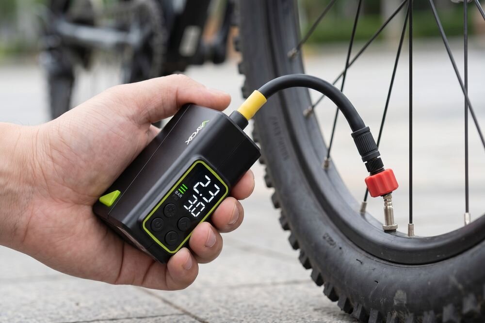 Osoba trzyma elektryczną pompkę rowerową z manometrem, podłączoną do wentyla opony rowerowej. Na wyświetlaczu widoczne są cyfry \'23\' i \'35\', a na urządzeniu znajduje się logo \'VAYOX\'.