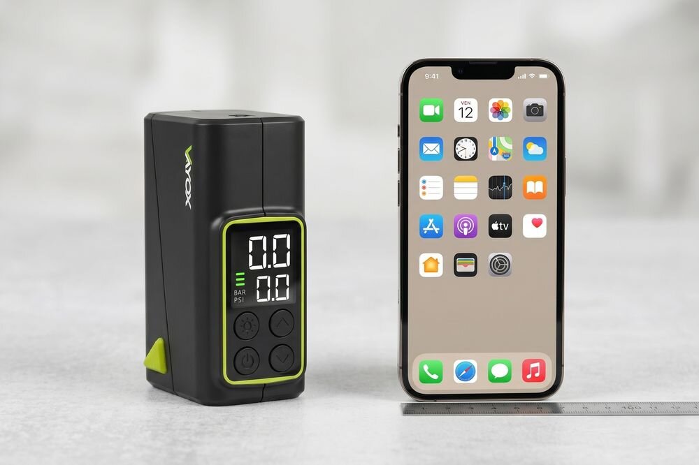 Na obrazie widoczna jest czarna pompka rowerowa z wyświetlaczem cyfrowym pokazującym \'0.0\' oraz smartfon z ekranem głównym pełnym ikon aplikacji. Obok znajduje się linijka, a na pompce widnieje logo \'VAYOX\'.