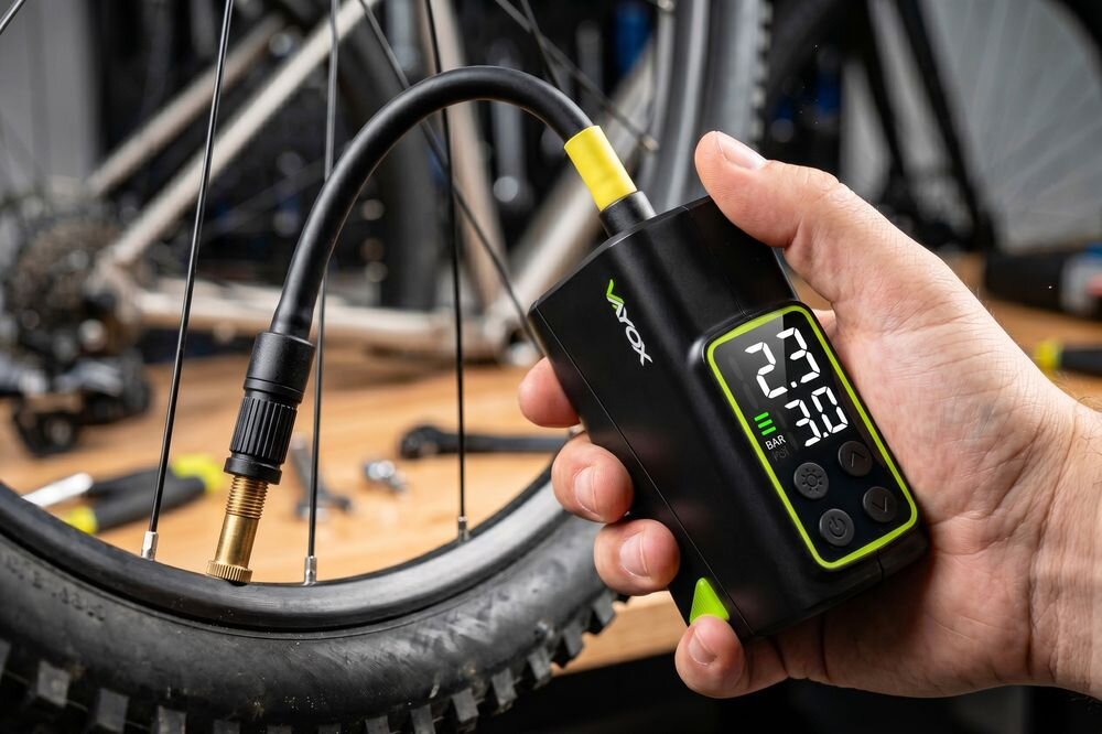 Osoba trzyma elektryczną pompkę rowerową z wyświetlaczem pokazującym wartości 23 i 3.0 BAR. Na urządzeniu widoczne jest logo \'VAYOX\'.