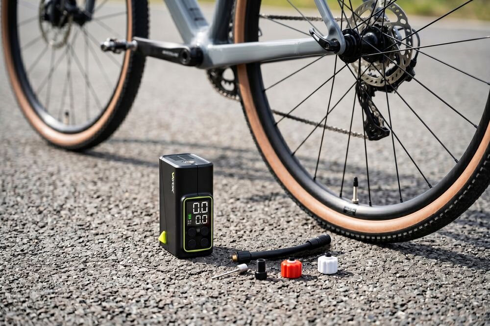 Na asfalcie stoi elektryczna pompka rowerowa z wyświetlaczem cyfrowym, obok której leżą różne końcówki. W tle widoczna jest część roweru z kołem.