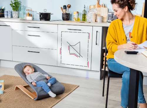 Kobieta siedzi przy stole w kuchni, obok niej na macie leży śpiące dziecko w leżaczku. Na szafce kuchennej widoczny jest symbol wyciszenia, sugerujący cichą pracę urządzenia.