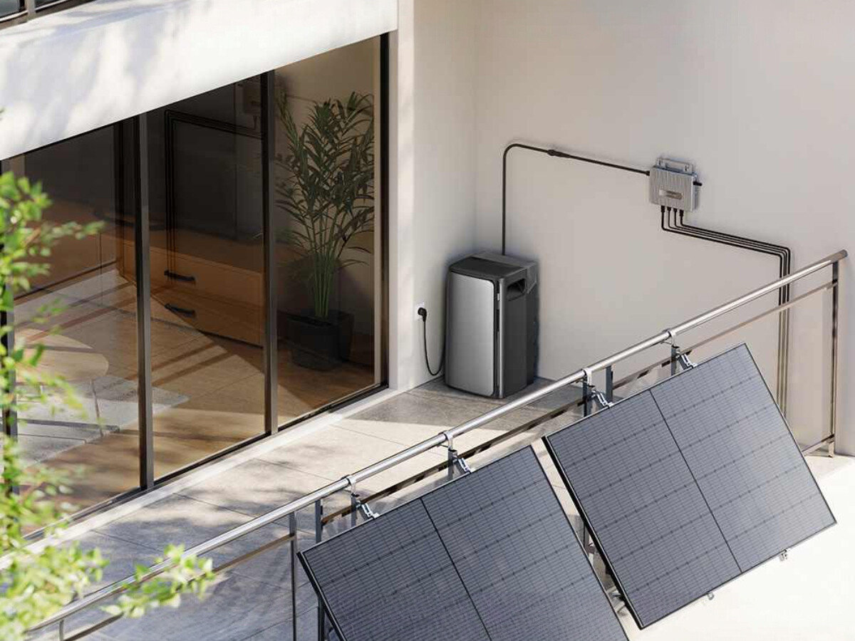 Na tarasie przed dużymi przeszklonymi drzwiami znajduje się urządzenie przypominające magazyn energii, obok którego widać instalację elektryczną. Na pierwszym planie widoczne są panele słoneczne zamontowane na balustradzie.