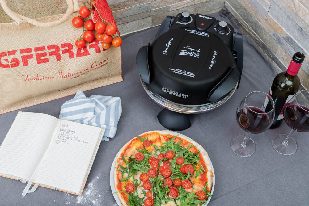 Na stole znajduje się czarny piec do pizzy z widocznym logo \'G3FERRARI\', obok leży pizza z rukolą i pomidorkami. W tle widać torbę z napisem \'G3FERRARI Tradizione Italiana in Cucina\', otwarty notes z przepisem oraz dwie lampki czerwonego wina i butelkę.