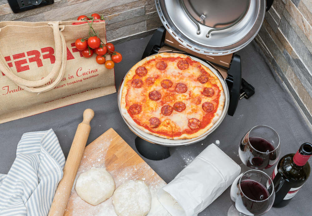 Na obrazie widoczny jest piec do pizzy z gotową pizzą z salami, obok leży deska z ciastem, wałek, kieliszki z czerwonym winem i torba z napisem \'FERRARI\'.