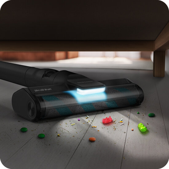 Odkurzacz z napisem \'Slim LED Brush\' odkurza podłogę pod meblem, oświetlając powierzchnię niebieskim światłem. Na podłodze widoczne są kolorowe okruchy i drobinki.