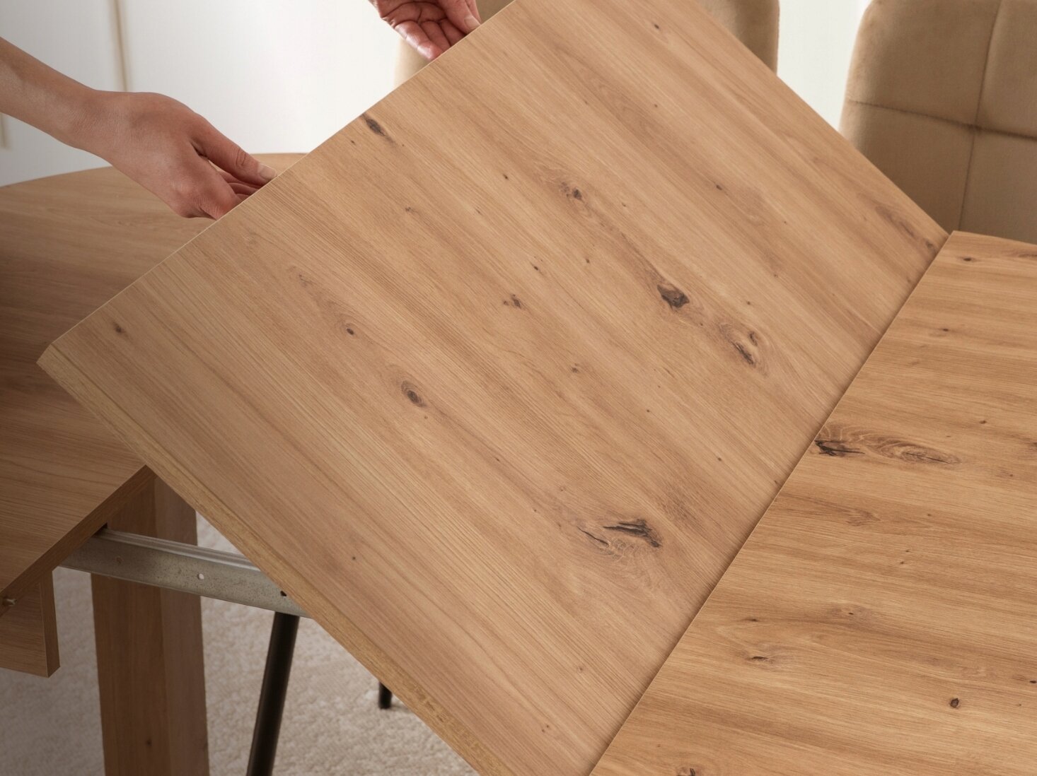 Osoba rozkłada drewniany stół, którego powierzchnia ma widoczne słoje i sęki. Stół znajduje się w jasnym pomieszczeniu z beżowymi krzesłami w tle.