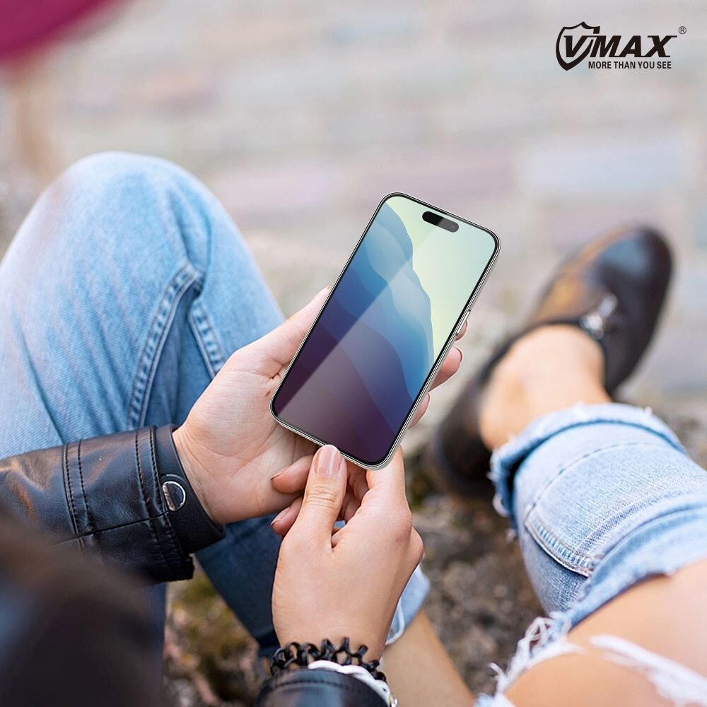 Osoba trzyma smartfon z widocznym szkłem ochronnym na ekranie, w tle rozmyte otoczenie. W prawym górnym rogu widoczne jest logo \'VMAX MORE THAN YOU SEE\'.