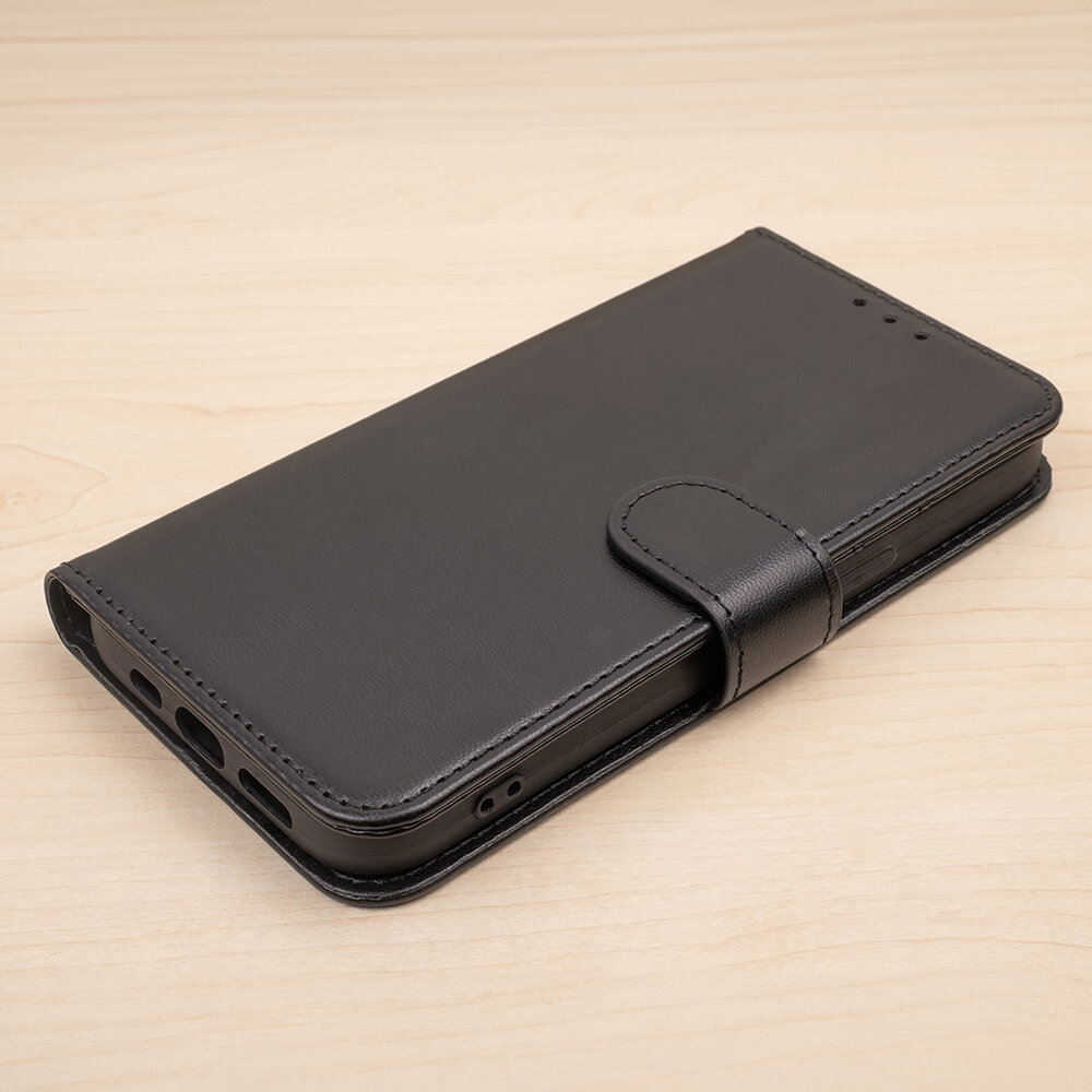 Czarne etui na telefon z klapką, wykonane z materiału imitującego skórę, leży na jasnym drewnianym tle. Etui posiada wycięcia na porty i przyciski oraz zapięcie na magnes.