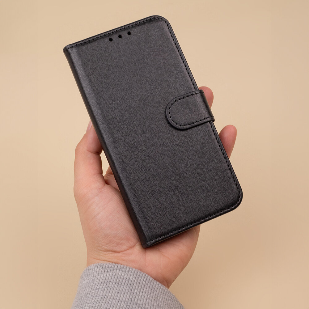 Osoba trzyma czarne etui na telefon z klapką, wykonane z materiału przypominającego skórę. Etui posiada zapięcie na magnes i otwory na głośnik.