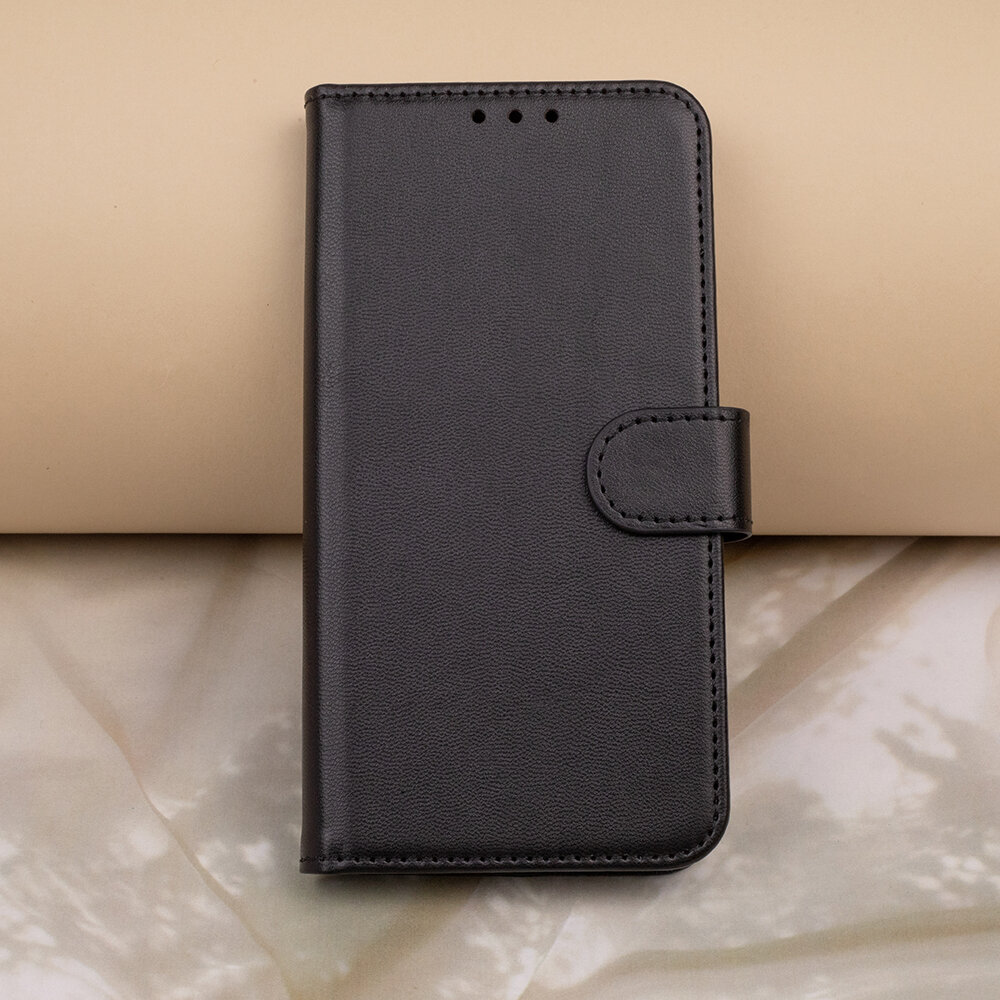 Czarne etui na telefon z klapką, wykonane z materiału imitującego skórę, z widocznym zapięciem na zatrzask. Tło jest w odcieniach beżu i szarości.