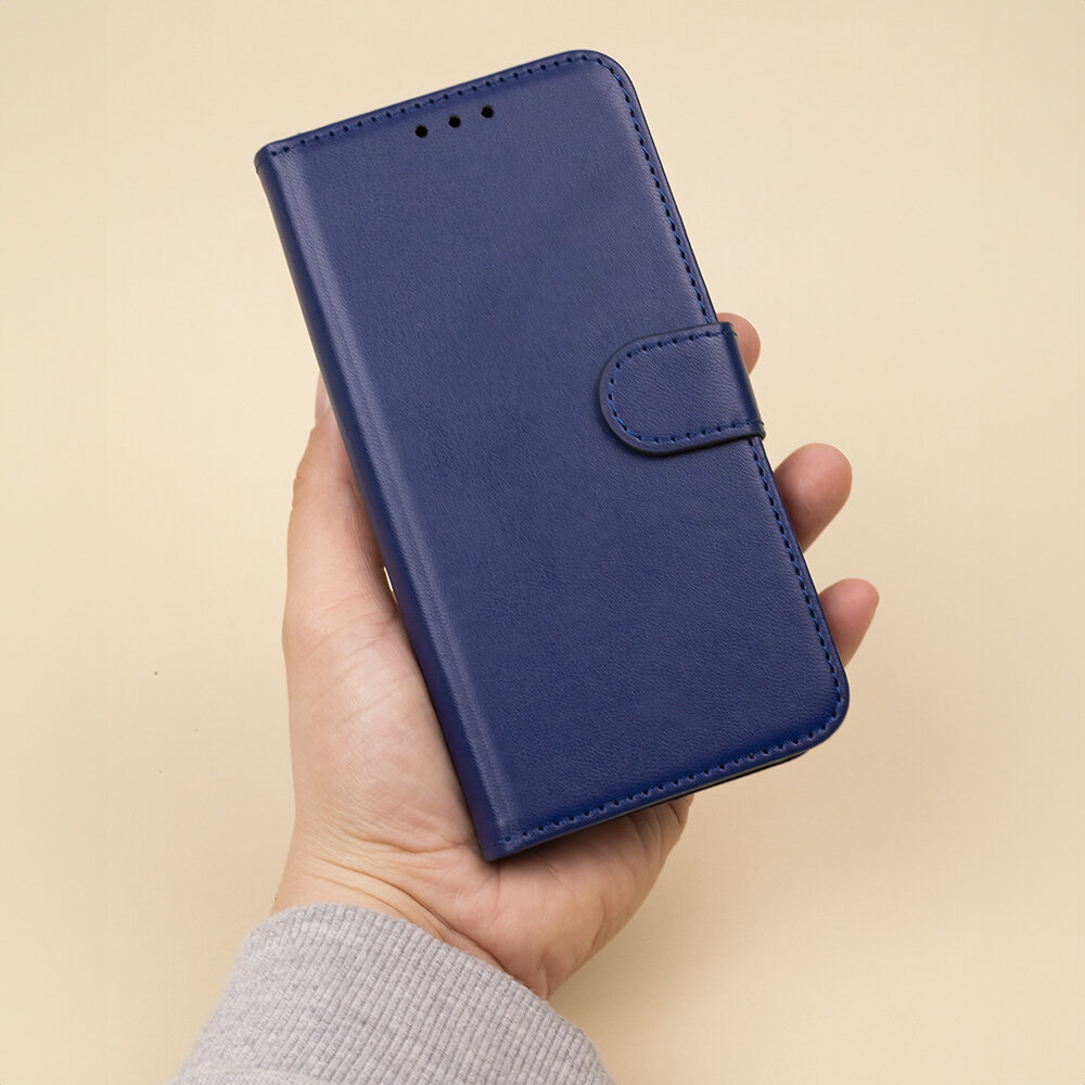 Osoba trzyma granatowe etui na telefon z klapką, wykonane z materiału przypominającego skórę. Etui posiada zapięcie na magnes i otwory na głośnik.