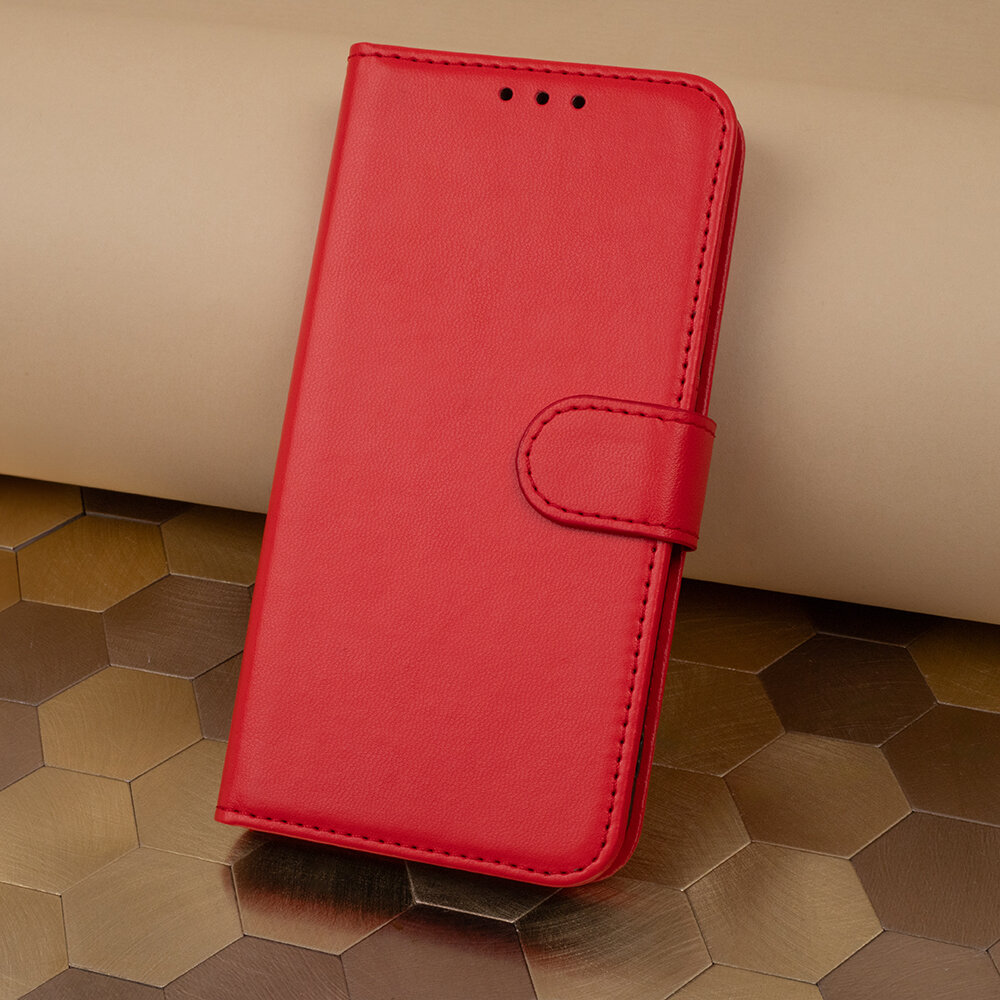 Czerwone etui na telefon z klapką, wykonane z materiału imitującego skórę, leży na powierzchni o wzorze heksagonalnym. Etui posiada zapięcie na magnes i trzy otwory na górze.