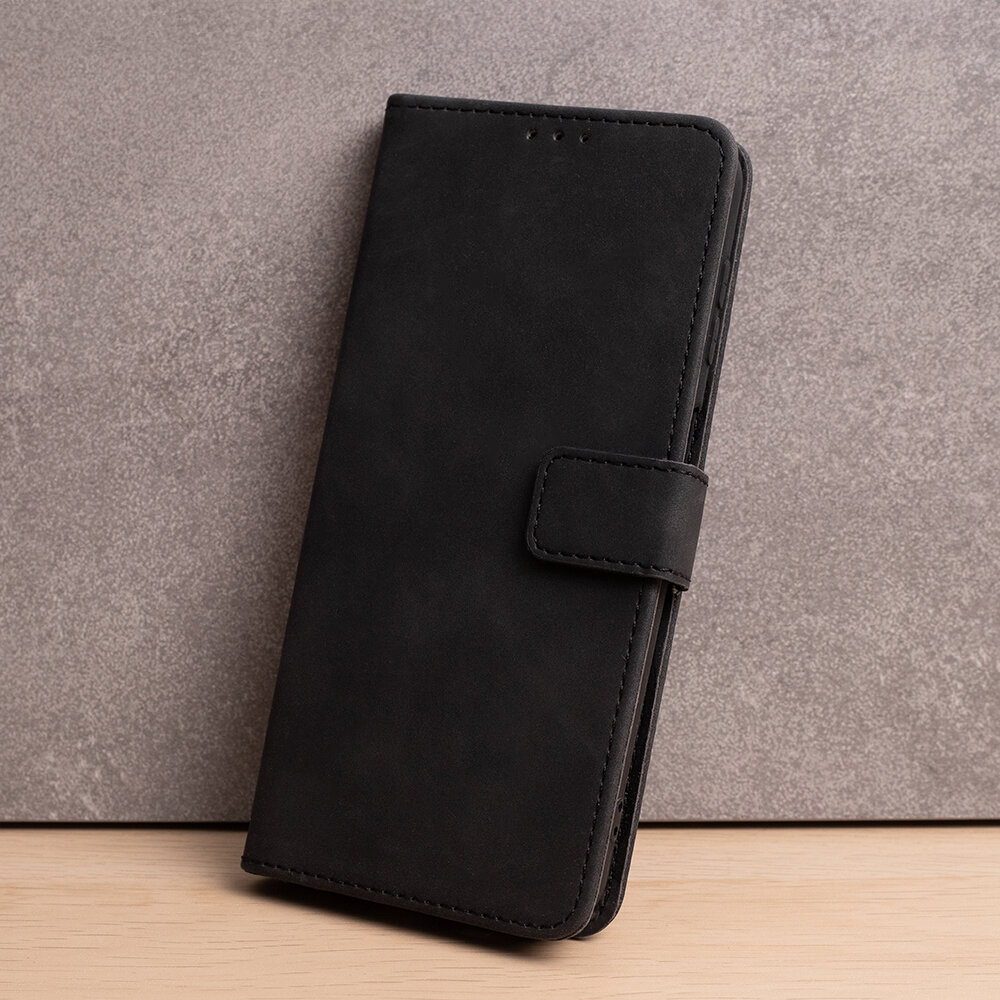 Czarne etui na telefon z klapką, wykonane z materiału przypominającego zamsz, stoi na tle szarej ściany i drewnianej powierzchni.
