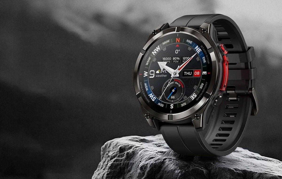 Czarny smartwatch z okrągłym wyświetlaczem, na którym widoczne są wskaźniki kierunków, data \'THU 08\' oraz różne parametry jak \'16000\', \'80%\', \'85\'. Urządzenie leży na kamiennej powierzchni, a tło jest rozmyte i ciemne.