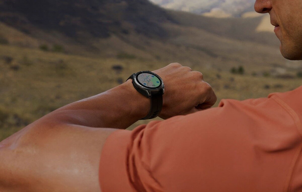 Osoba w pomarańczowej koszulce patrzy na czarny smartwatch na nadgarstku, na tle rozmytego krajobrazu.