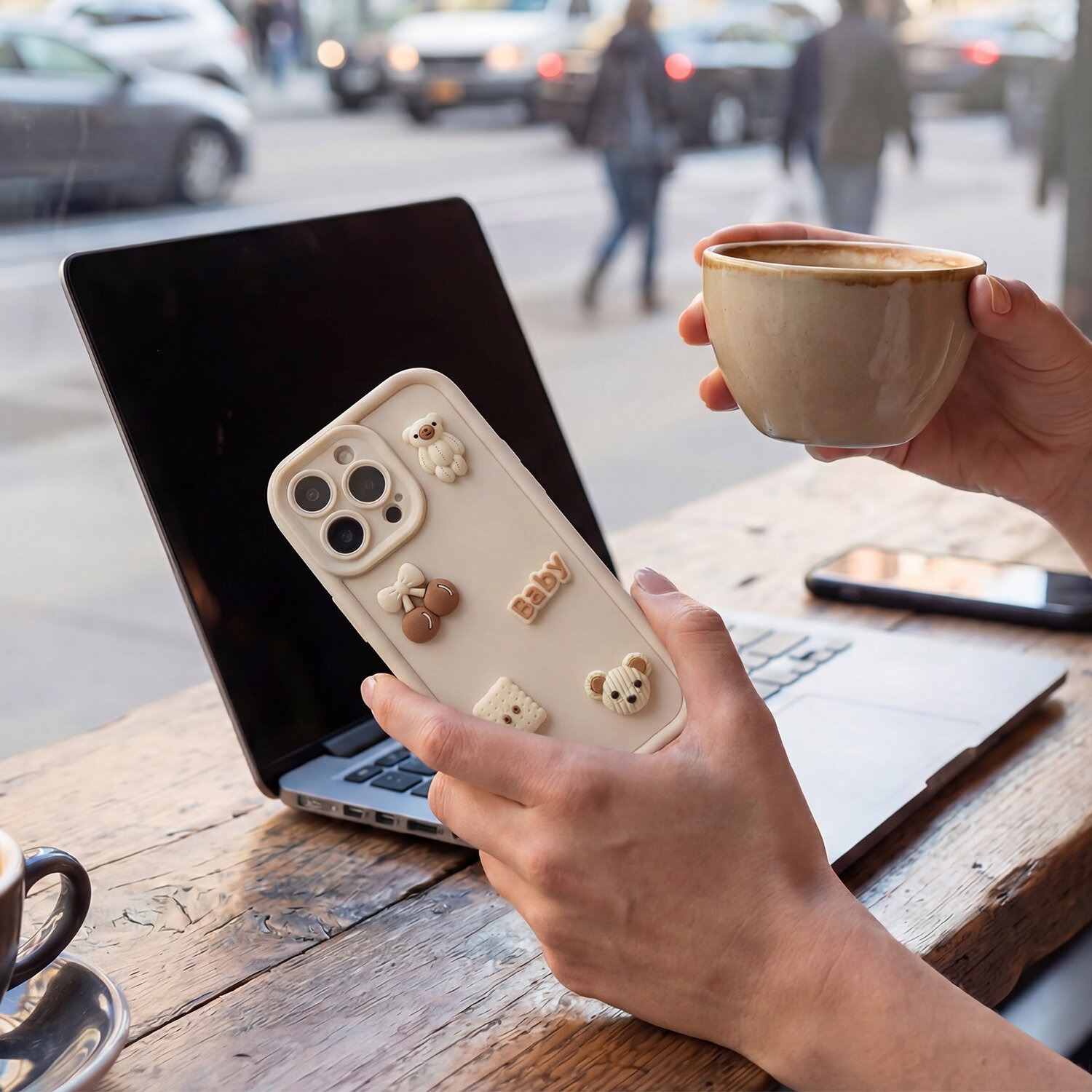 Osoba trzyma telefon z nakładką ozdobioną małymi figurkami misiów i napisem \'baby\', w tle widać laptopa i filiżankę z napojem.