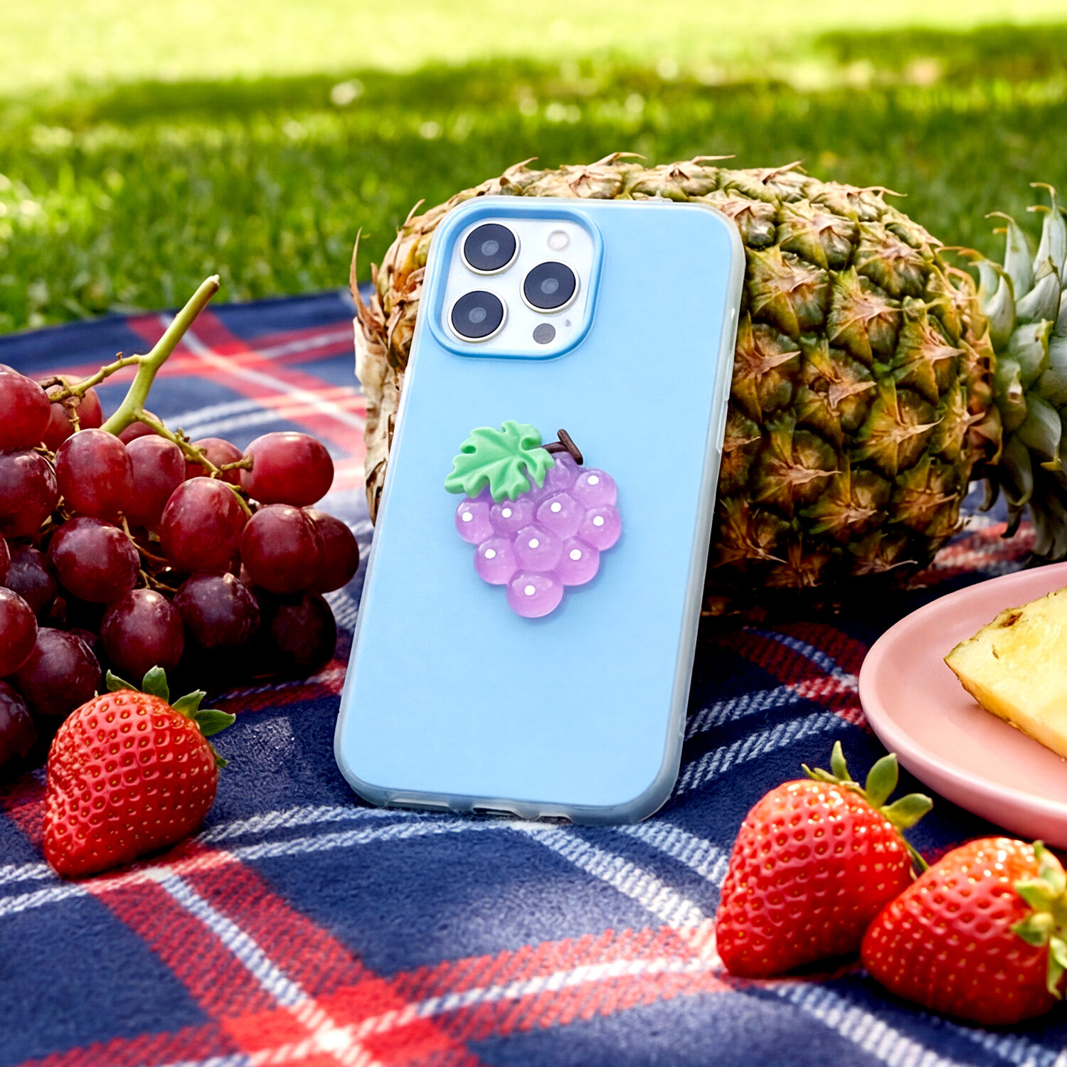 Na kocu w kratę leży niebieska nakładka na telefon z motywem winogron. W tle widoczne są owoce: ananas, winogrona i truskawki.