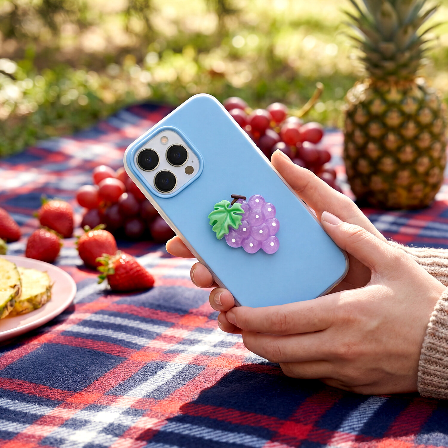 Osoba trzyma telefon z niebieską nakładką ozdobioną fioletowym winogronem. W tle widać owoce, takie jak truskawki, winogrona i ananas, na kocu w kratę.