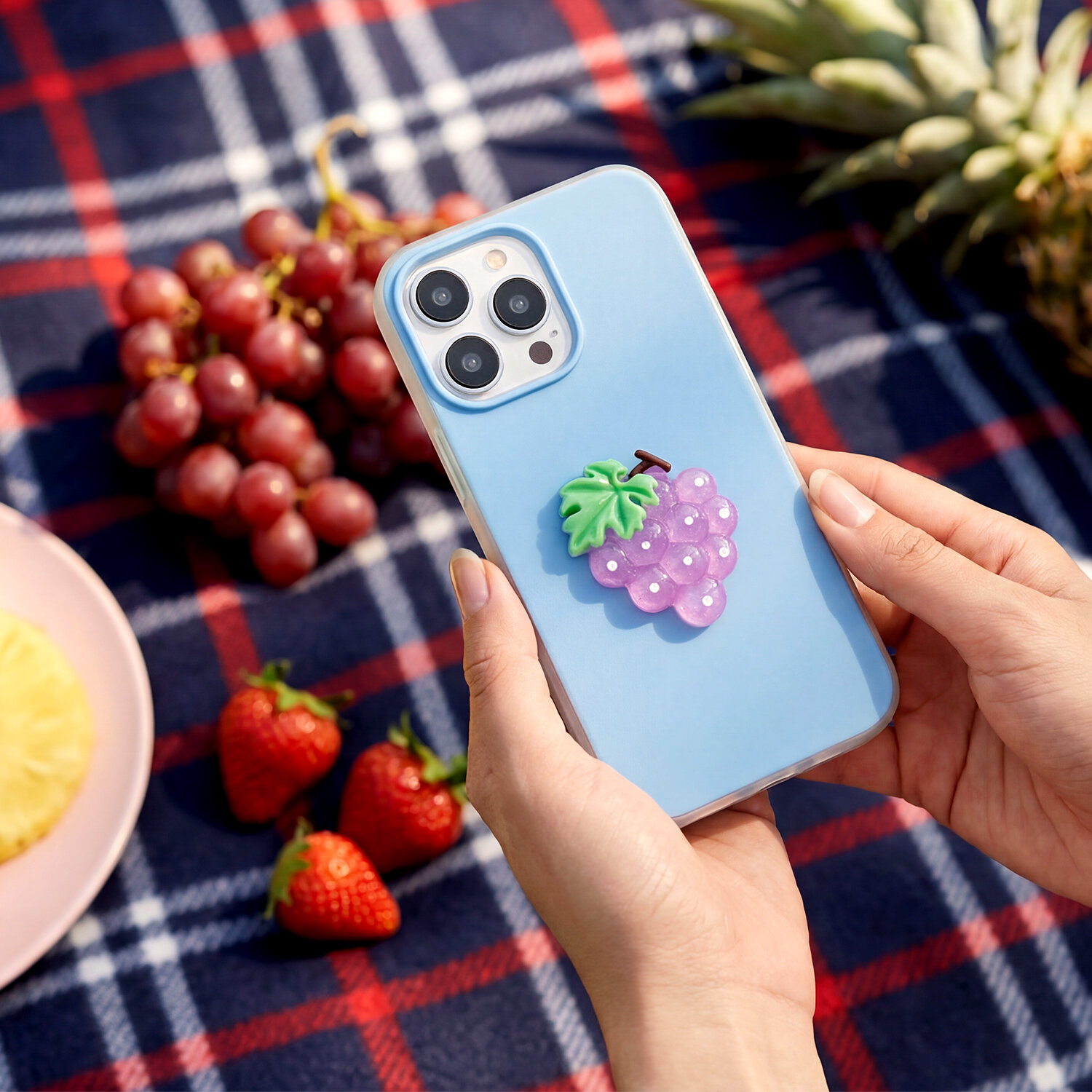 Osoba trzyma telefon z niebieską nakładką ozdobioną fioletowym winogronem. W tle widać owoce, takie jak winogrona, truskawki i ananas, na kraciastej tkaninie.