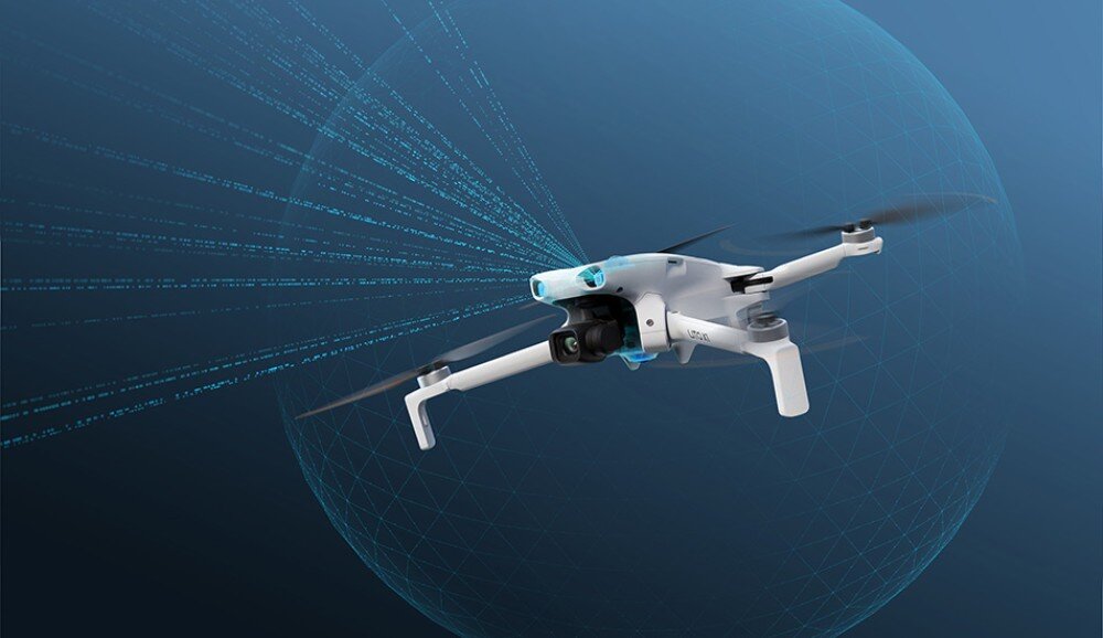 Na obrazie widoczny jest biały dron z czterema śmigłami, unoszący się w powietrzu na tle niebieskiego, geometrycznego wzoru. Dron ma zamontowaną kamerę z przodu.