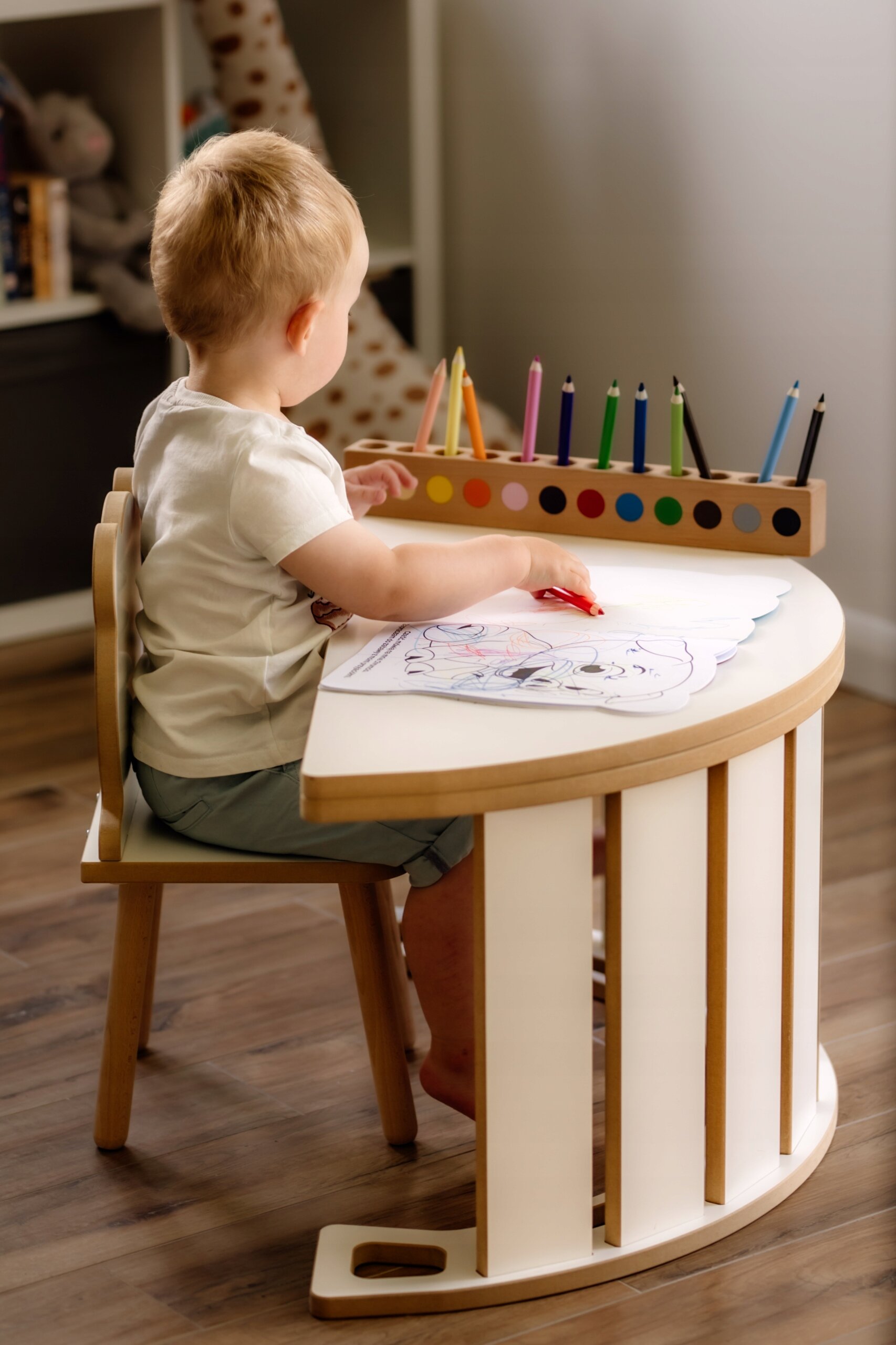 Dziecko siedzi przy drewnianym stoliku z kolorowymi kredkami, rysując na kartce papieru. W tle widać półkę z pluszowymi zabawkami.
