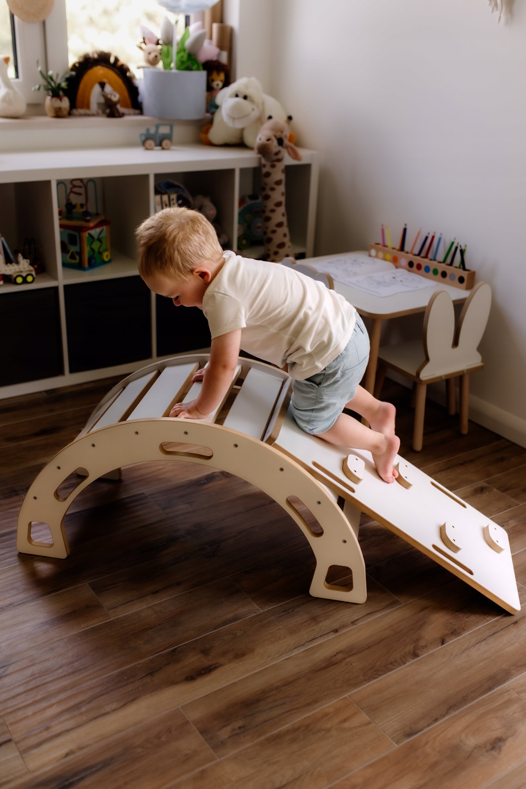 Dziecko bawi się na białym bujaku Montessori w kształcie mostka, znajdującym się w pokoju dziecięcym z drewnianą podłogą. W tle widać półki z zabawkami i stolik z krzesełkami.