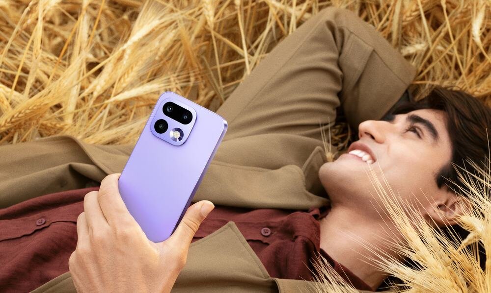 Osoba leży na polu zboża, trzymając w ręku fioletowy smartfon z trzema aparatami na tylnej obudowie.