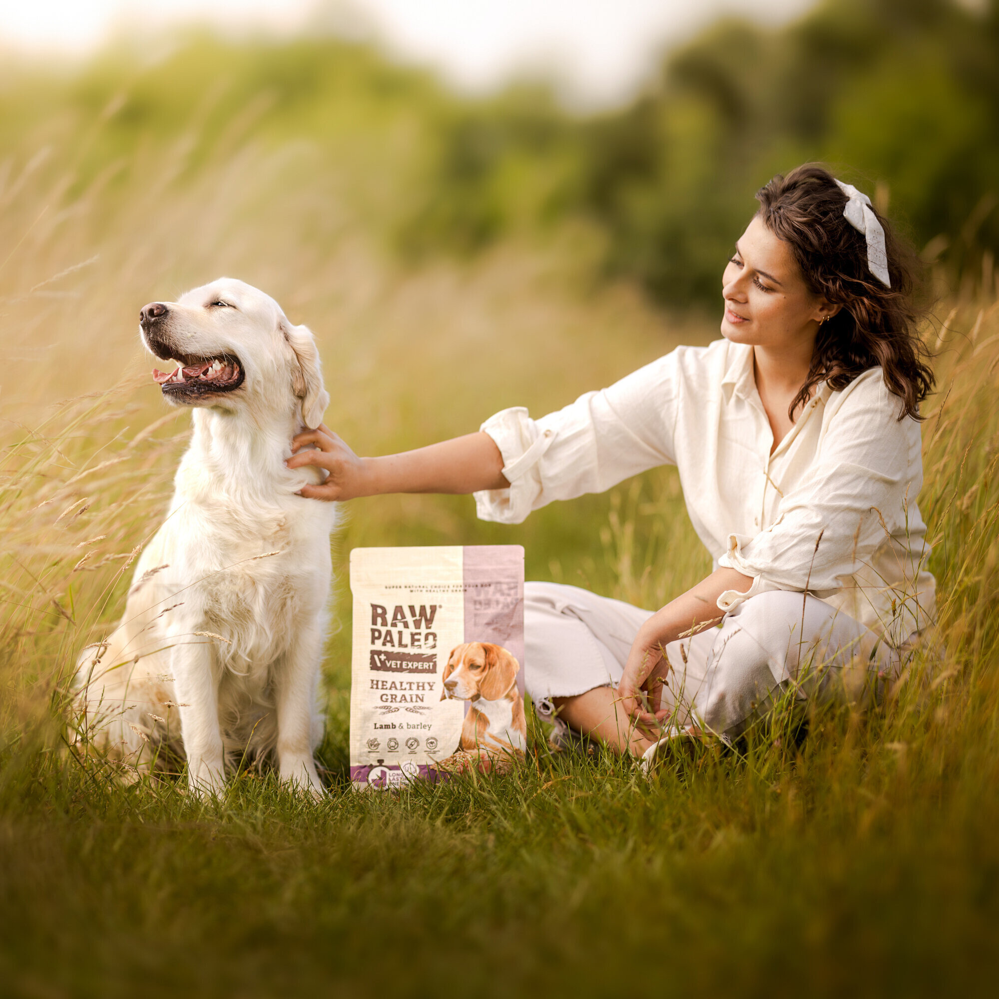 Na obrazie widać psa siedzącego na trawie obok osoby, która go głaszcze. Obok znajduje się opakowanie karmy z napisem \'RAW PALEO VET EXPERT HEALTHY GRAIN Lamb & Barley\'.