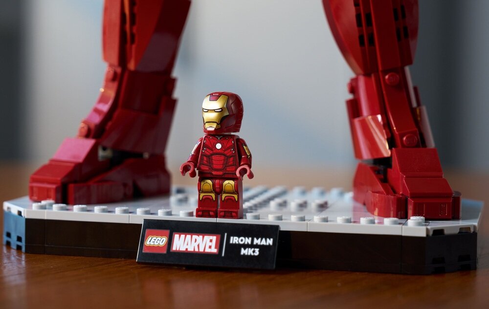 Na obrazie znajduje się figurka LEGO przedstawiająca Iron Mana w czerwono-złotej zbroi, stojąca na podstawce. Na podstawce widoczna jest tabliczka z napisem \'LEGO MARVEL IRON MAN MK3\'.