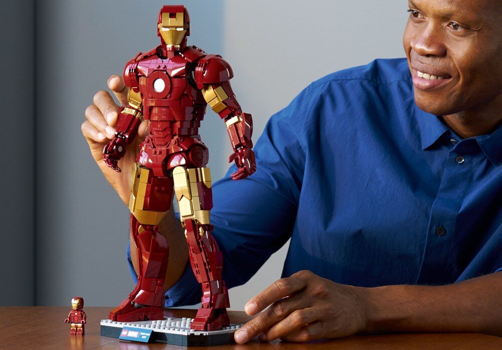 Na stole stoi duża figurka Iron Mana z klocków LEGO oraz mniejsza wersja tej samej postaci. Obok znajduje się osoba w niebieskiej koszuli, która patrzy na figurkę.