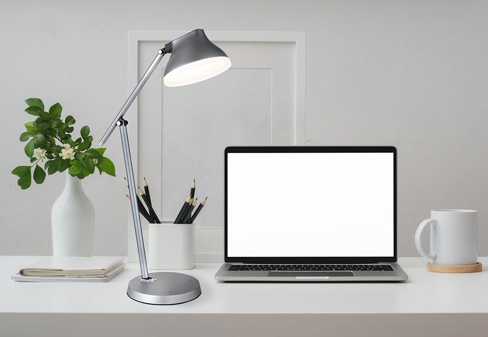 Na biurku znajduje się szara lampa biurkowa LED, laptop z pustym ekranem, biały kubek oraz wazon z zieloną rośliną. W tle widać ramkę na zdjęcia bez zdjęcia.