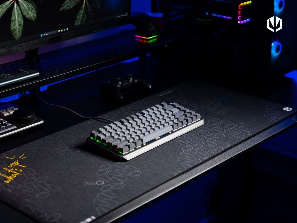 Na biurku znajduje się klawiatura mechaniczna z podświetleniem RGB, umieszczona na dużej podkładce z geometrycznym wzorem. W tle widoczny jest monitor z roślinną tapetą oraz komputer z podświetleniem LED.