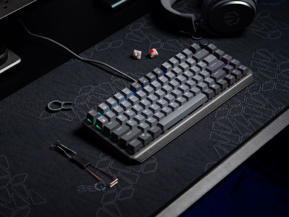 Na ciemnym biurku znajduje się klawiatura mechaniczna z podświetleniem RGB. Obok leżą słuchawki, kilka przełączników i narzędzie do wyjmowania klawiszy.
