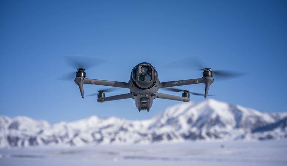 Dron w locie na tle ośnieżonych gór i błękitnego nieba. Na dronie widoczny jest napis \'Mavic 4 Pro\'.