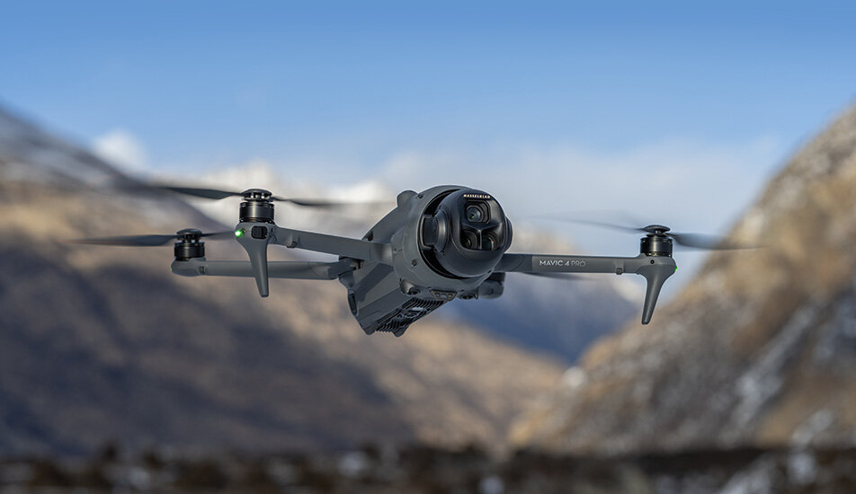 Dron w locie na tle górskiego krajobrazu, z widocznym napisem \'MAVIC 2 PRO\' na ramieniu. Dron ma cztery śmigła i kamerę z przodu.