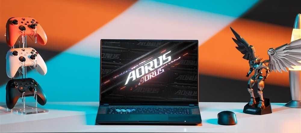 Na biurku znajduje się laptop z wyświetlonym logo \'AORUS 30URS\'. Obok laptopa stoją trzy kontrolery do gier na stojaku oraz figurka skrzydlatego robota z napisem \'AORUS\' na podstawce.