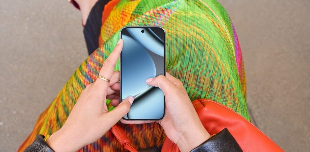 Osoba trzyma smartfon z wyświetlaczem włączonym, na którym widoczny jest abstrakcyjny wzór. W tle znajduje się kolorowa tkanina z wzorem w zielone, pomarańczowe i żółte pasy.