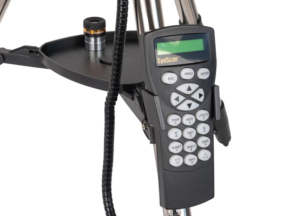 Na obrazie widoczny jest panel sterowania SynScan z wyświetlaczem i przyciskami, zamocowany na statywie teleskopu. Obok znajduje się okular teleskopu umieszczony na półce.