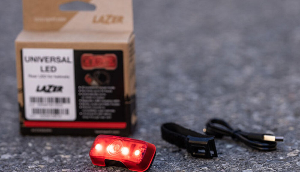 Na obrazie widoczna jest czerwona lampka LED, opakowanie z napisem \'UNIVERSAL LED\' oraz kabel USB. Logo \'LAZER\' znajduje się na opakowaniu.