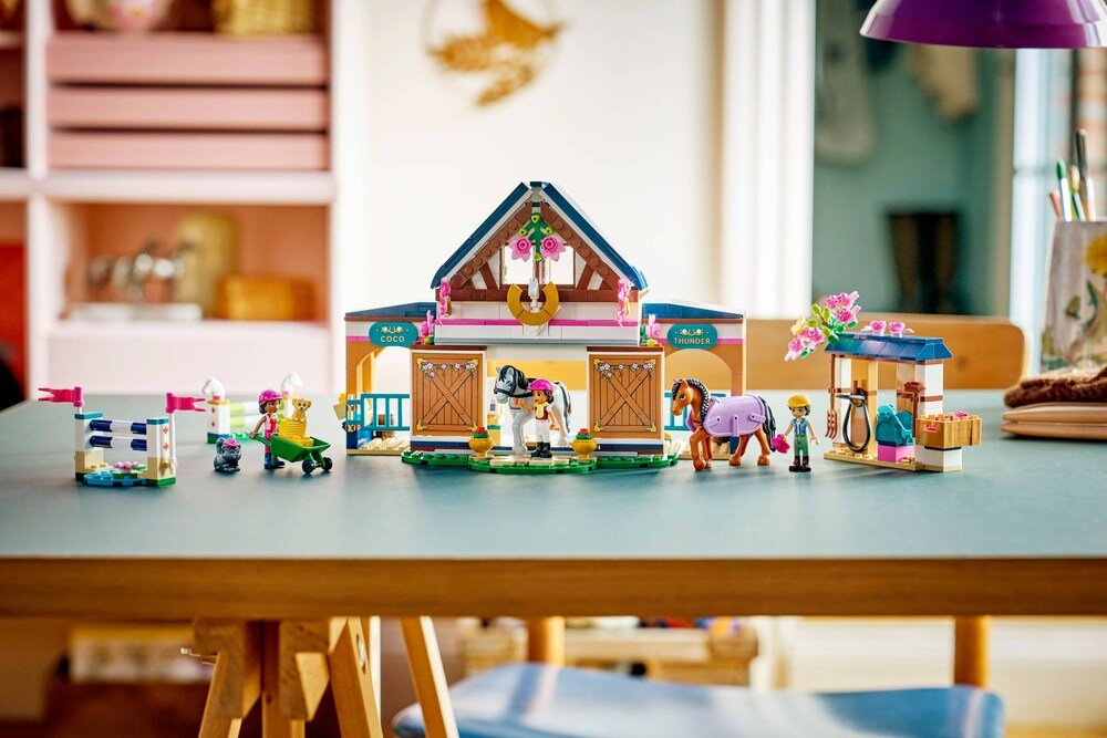 Na stole znajduje się zestaw LEGO przedstawiający stadninę i szkółkę jeździecką z budynkiem, końmi i figurkami. Widoczne są detale takie jak kwiaty, płotki i akcesoria jeździeckie.