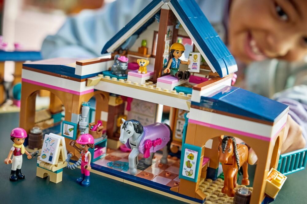 Zestaw LEGO przedstawiający stadninę i szkółkę jeździecką z figurkami koni i postaci. W tle widoczna jest osoba bawiąca się zestawem.
