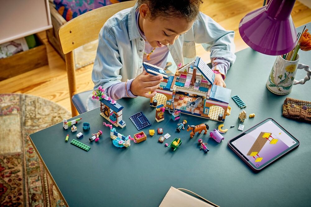Na stole znajduje się zestaw klocków LEGO przedstawiający budynek i różne akcesoria, obok leży tablet z instrukcją. Osoba bawi się klockami, a w tle widać lampkę i kubek z kwiatami.