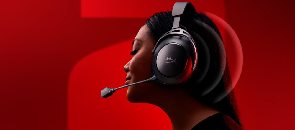 Osoba nosi nauszne słuchawki z mikrofonem na tle czerwonej ściany. Na słuchawkach widoczne jest logo HyperX.