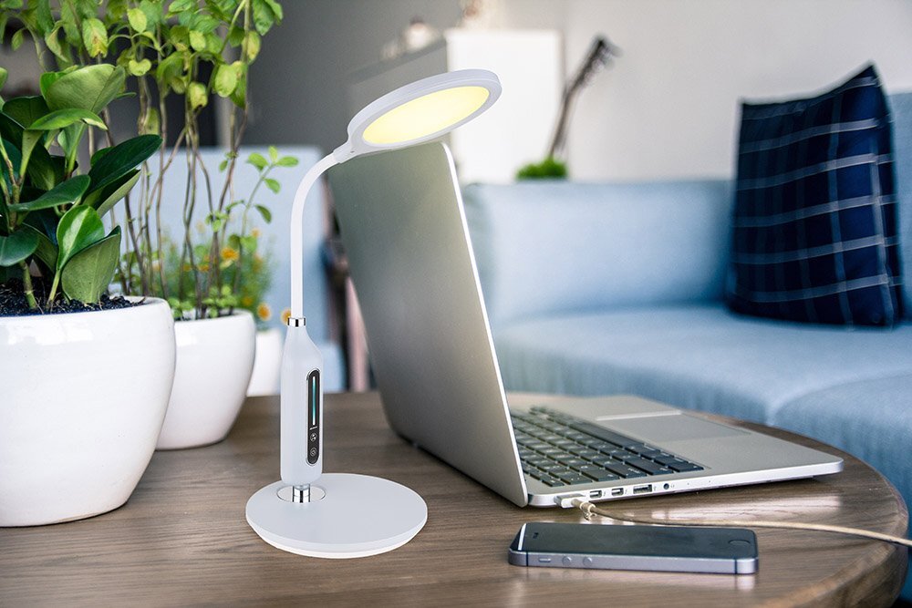 Na drewnianym stole znajduje się biała, dotykowa lampa biurkowa LED obok otwartego laptopa i smartfona. W tle widoczna jest kanapa z poduszką oraz rośliny doniczkowe.