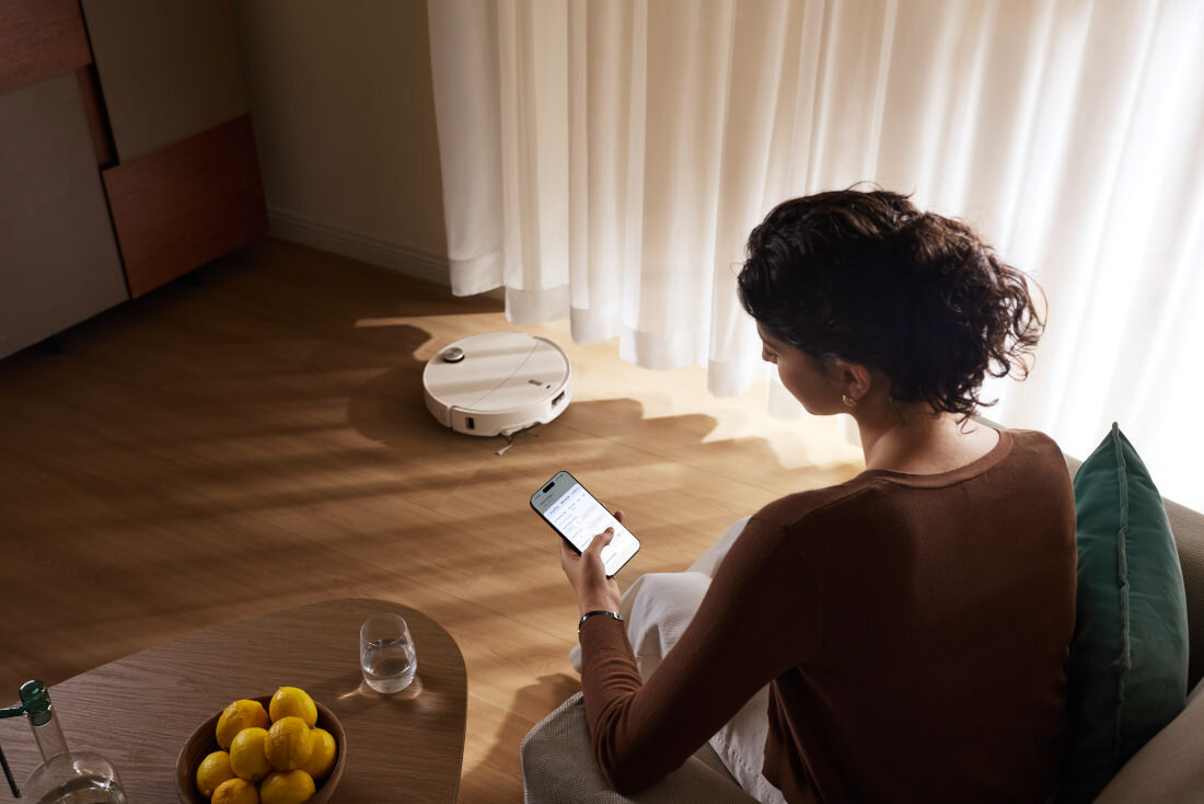 Osoba siedzi na kanapie, trzymając smartfon, podczas gdy biały robot sprzątający porusza się po podłodze. Na stoliku obok znajduje się miska z cytrynami i szklanka wody.