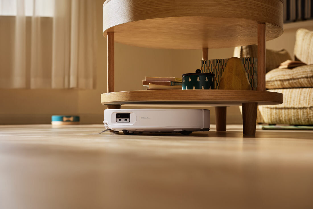 Biały robot sprzątający znajduje się pod drewnianym stolikiem w jasnym pomieszczeniu. Na stoliku leżą książki i ozdobne pudełko.