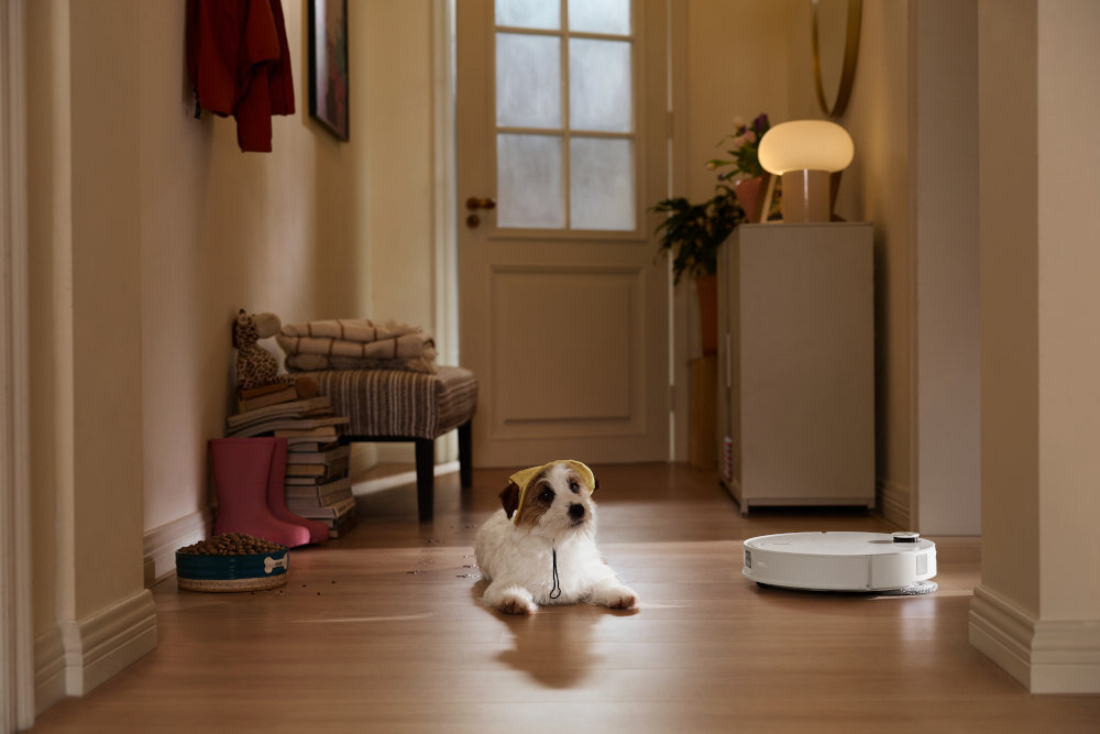 W korytarzu na drewnianej podłodze leży mały pies, obok którego znajduje się biały robot sprzątający. W tle widać szafkę z lampą i stos książek na podłodze.